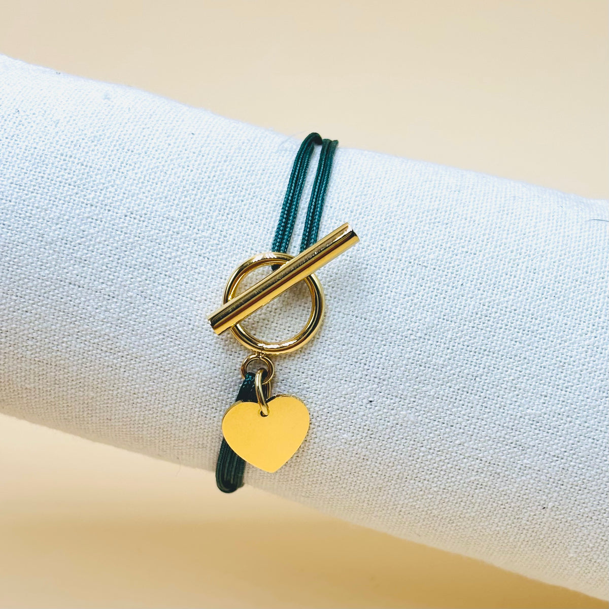 Bracelet cordon vert foncé avec fermoir T doré et cœur à graver, sur un présentoir en tissu.