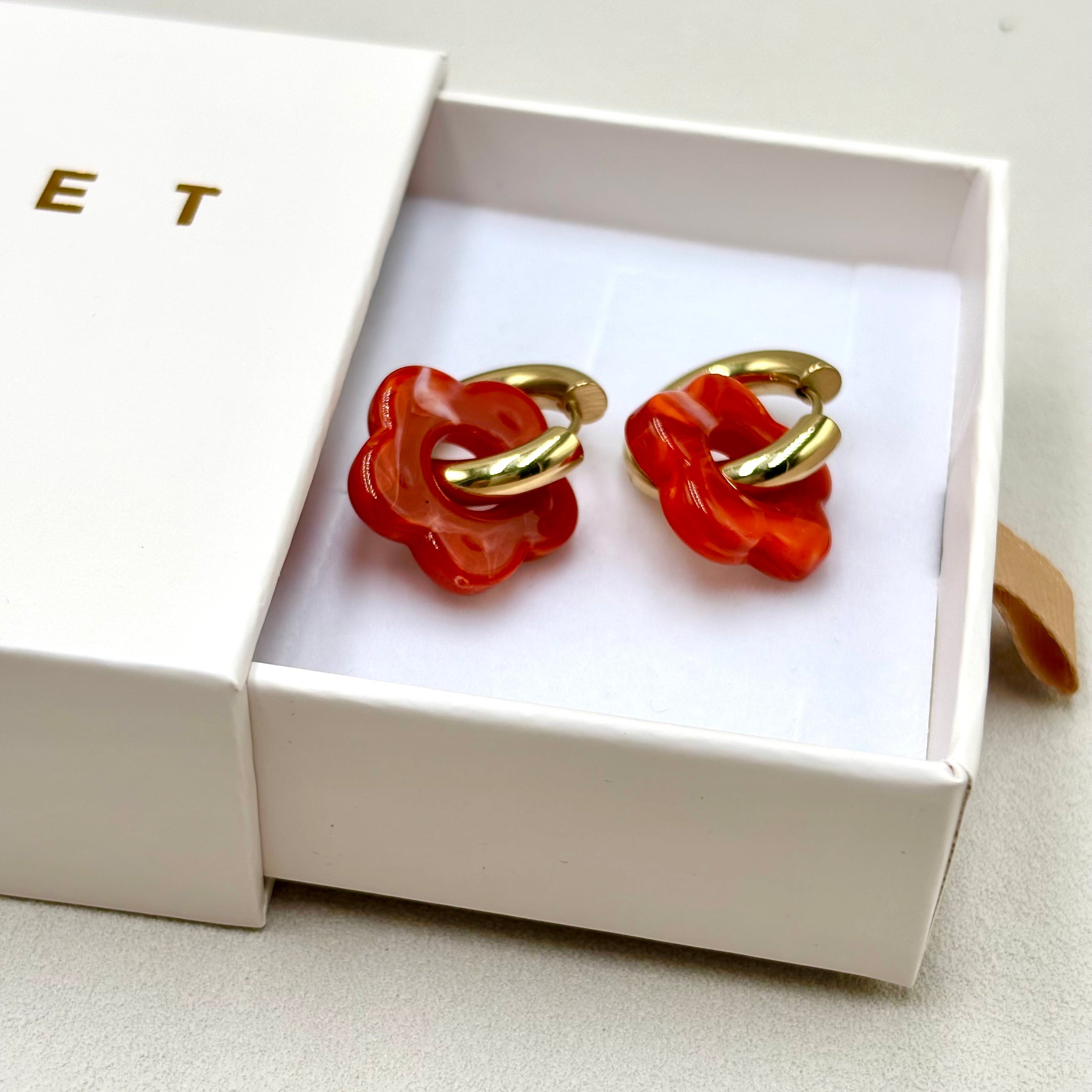 Boucles d'oreilles créoles en acier doré orné d'une fleur en acétate rouge orangé présentées dans une boite cadeau