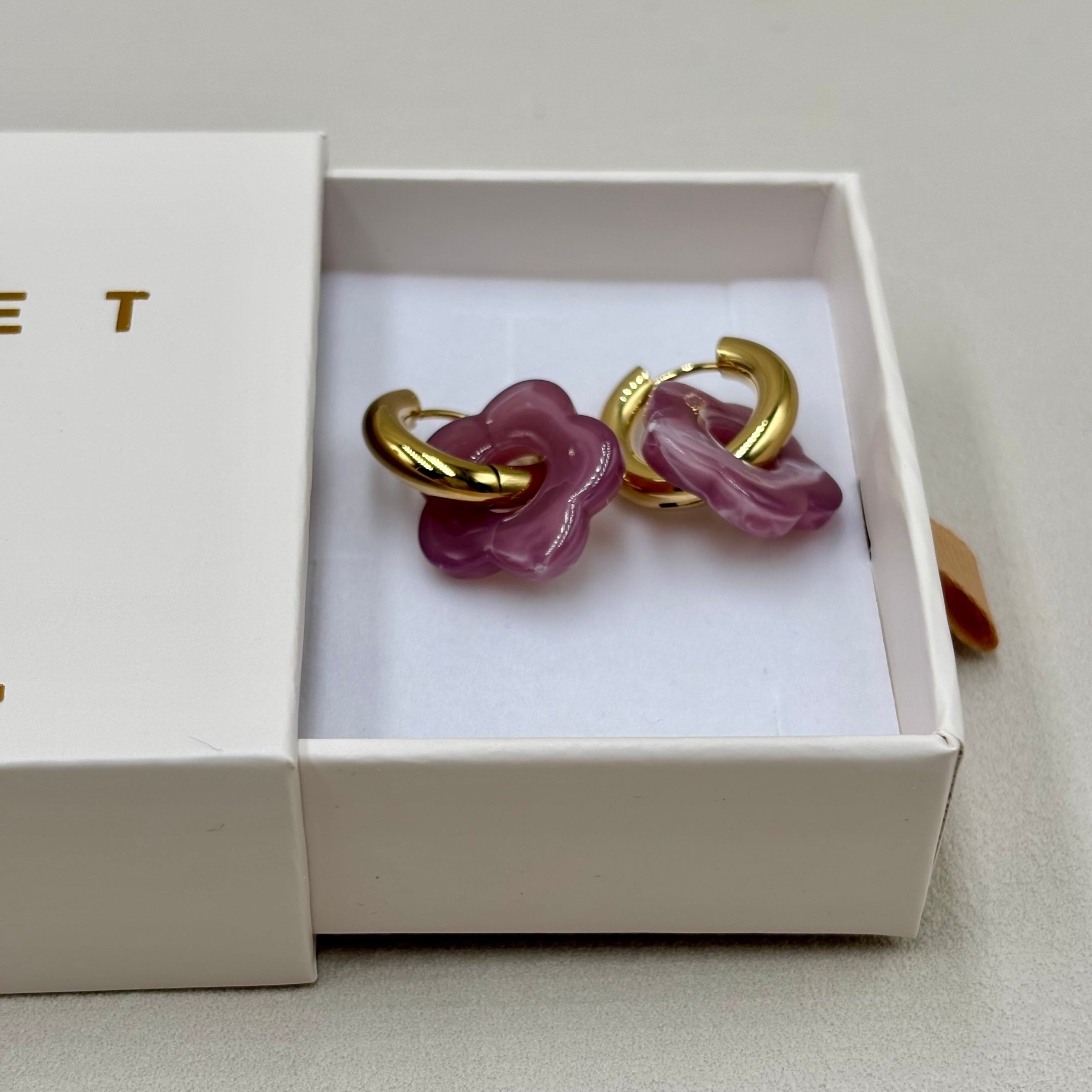 Boucles d'oreilles créoles en acier doré orné d'une fleur en acétate violet présentées dans une boite cadeau
