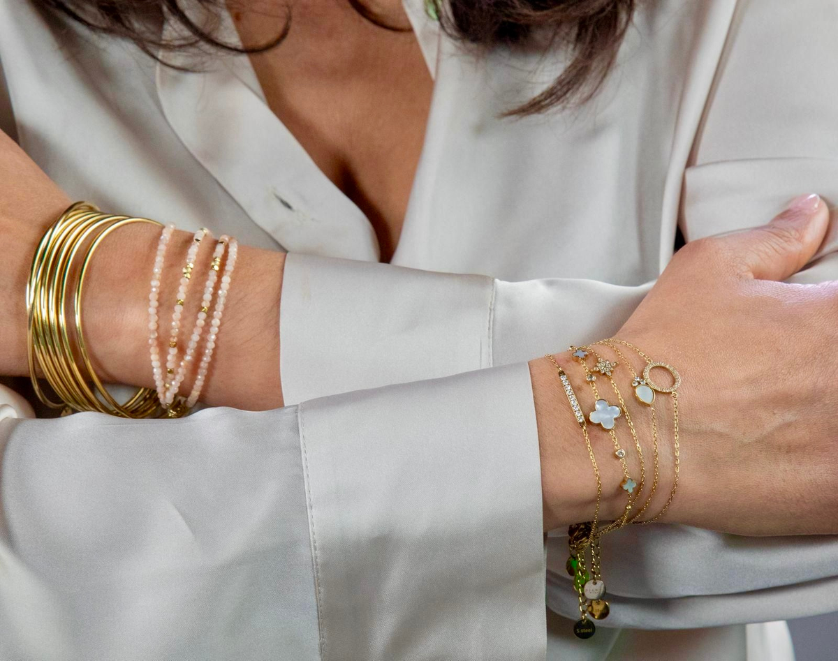 Bracelets fins dorés en acier inoxydable motifs en nacre et en zirconium portés avec un chemisier satin beige clair