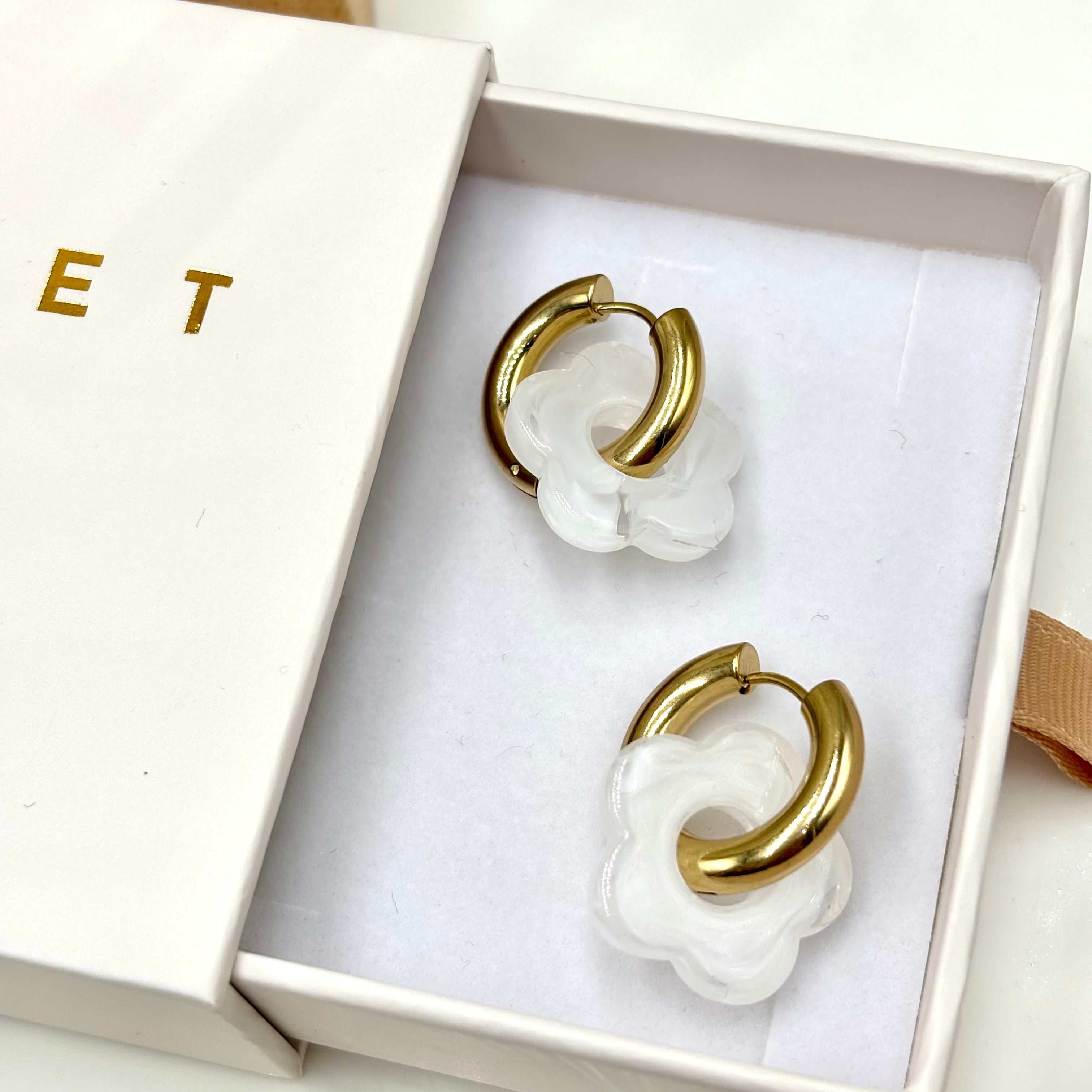 Boucles d'oreilles créoles en acier doré orné d'une fleur en acétate blanc présentées dans une boite cadeau Djanet Bijoux