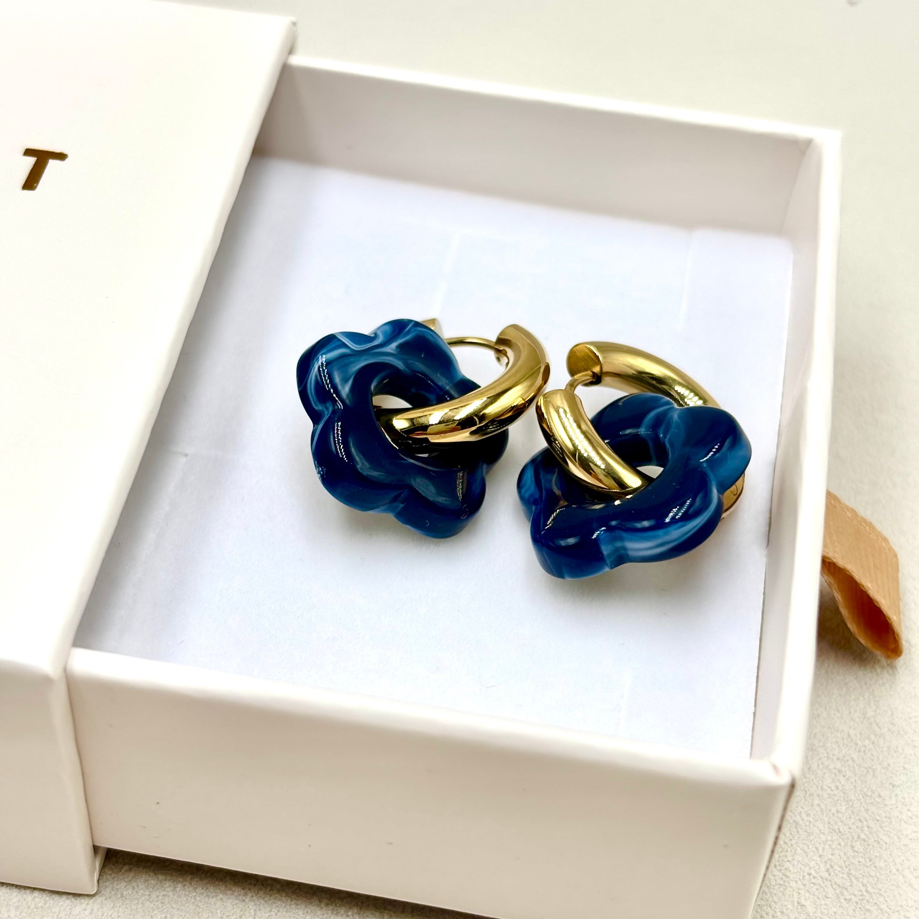 Boucles d'oreilles créoles en acier doré orné d'une fleur en acétate bleu foncé présentées dans une boite cadeau 
