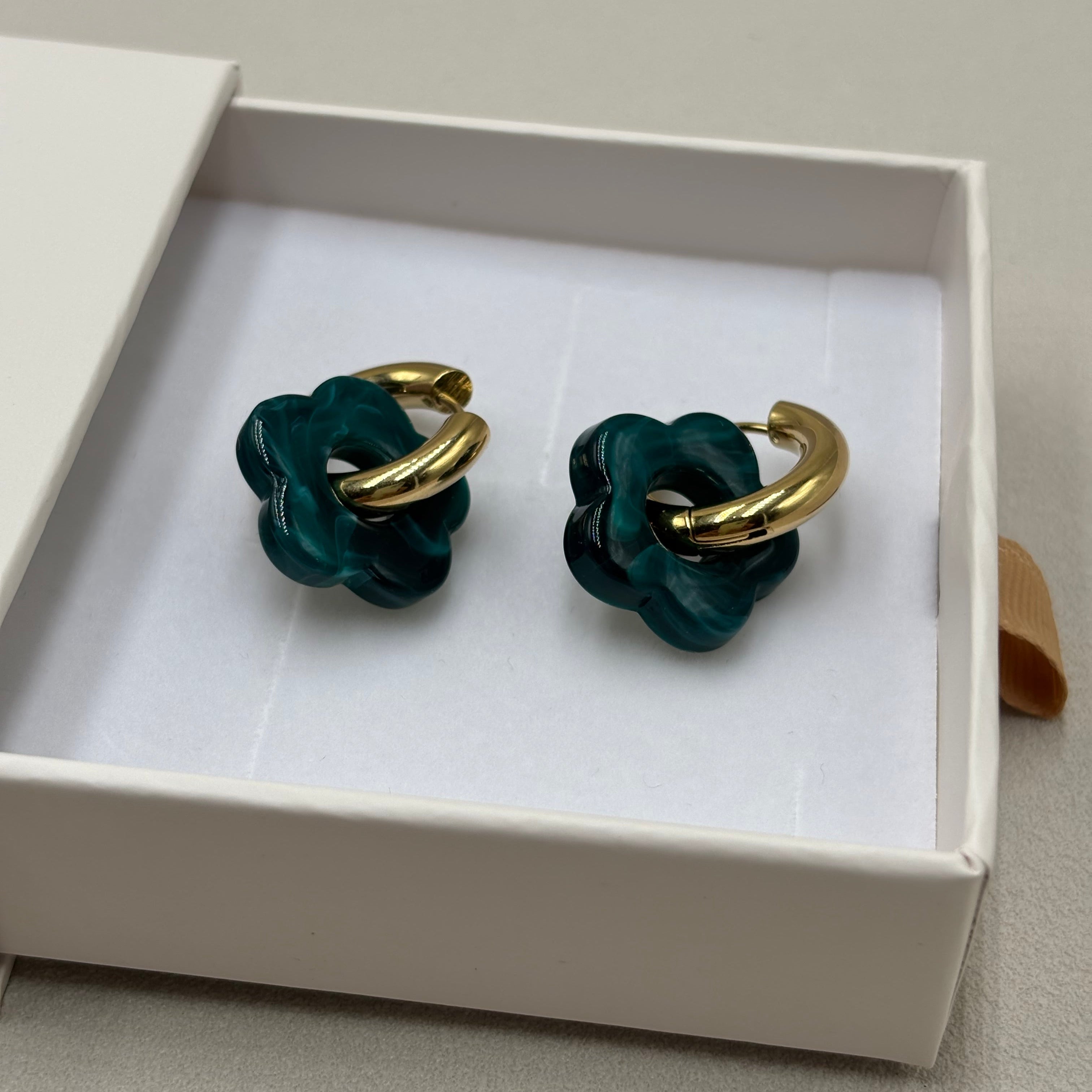 Boucles d'oreilles créoles en acier doré orné d'une fleur en acétate bleu pétrole présentées dans une boite cadeau