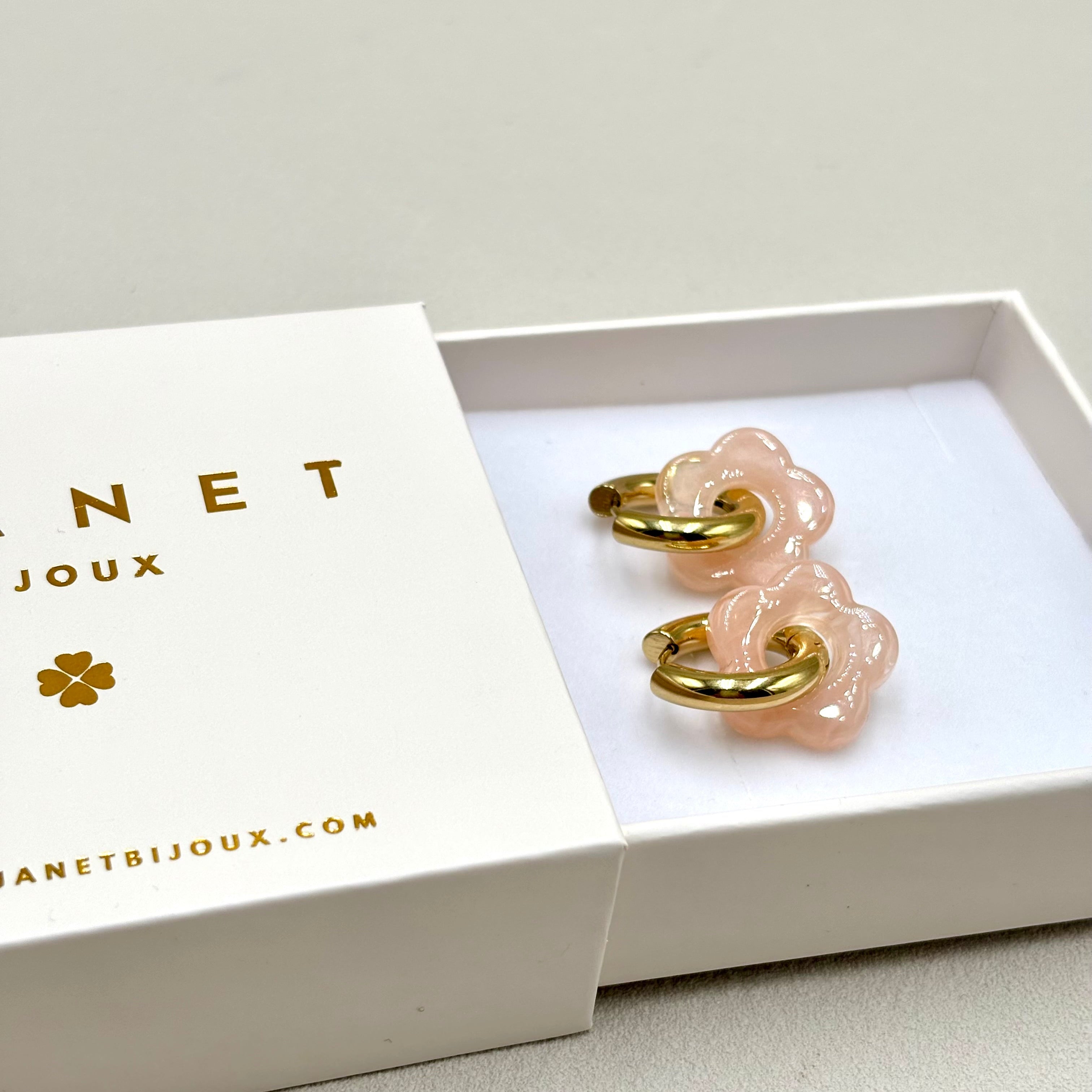 Boucles d'oreilles créoles en acier doré orné d'une fleur en acétate rose clair présentées dans une boite cadeau Djanet Bijoux